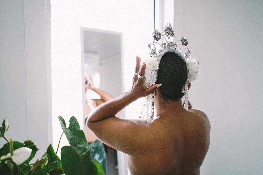 A topless drag queen putting on a crown in a mirror with green plants in background.