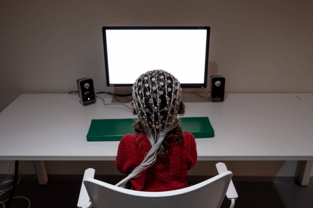 Back view of little girl in EEG cap looking at computer recording electrical stimuli of encephalogram and eye tracking during research