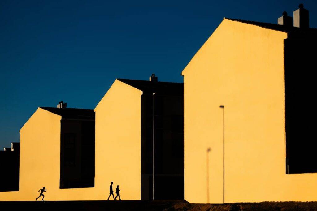 Photograph of a street with three houses with yellow facades in a minimalist key and three silhouettes walking along the sidewalk