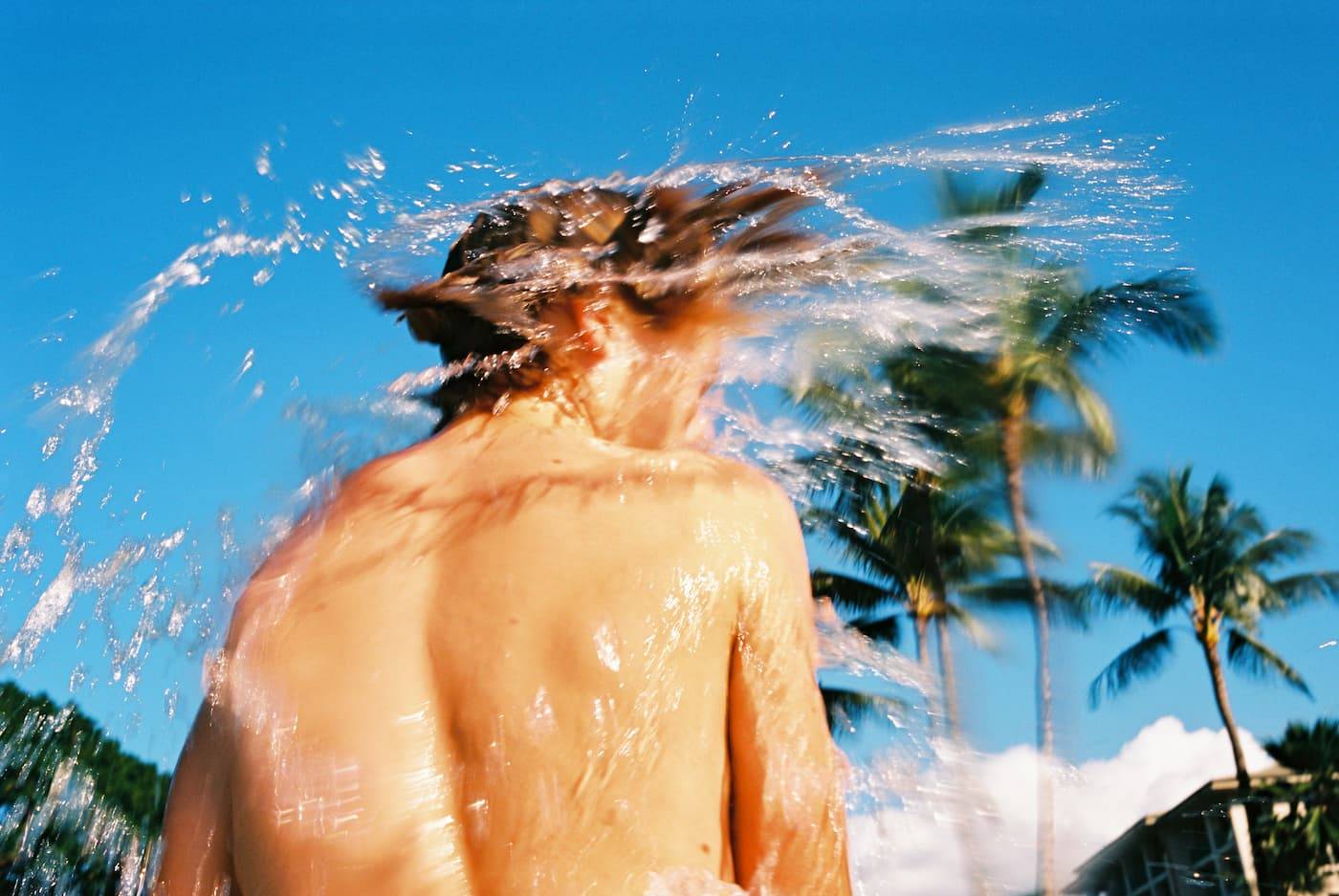 Boy in Surf shaking his wet hair and splashing water, vivid colors, plam trees and blue sky in background