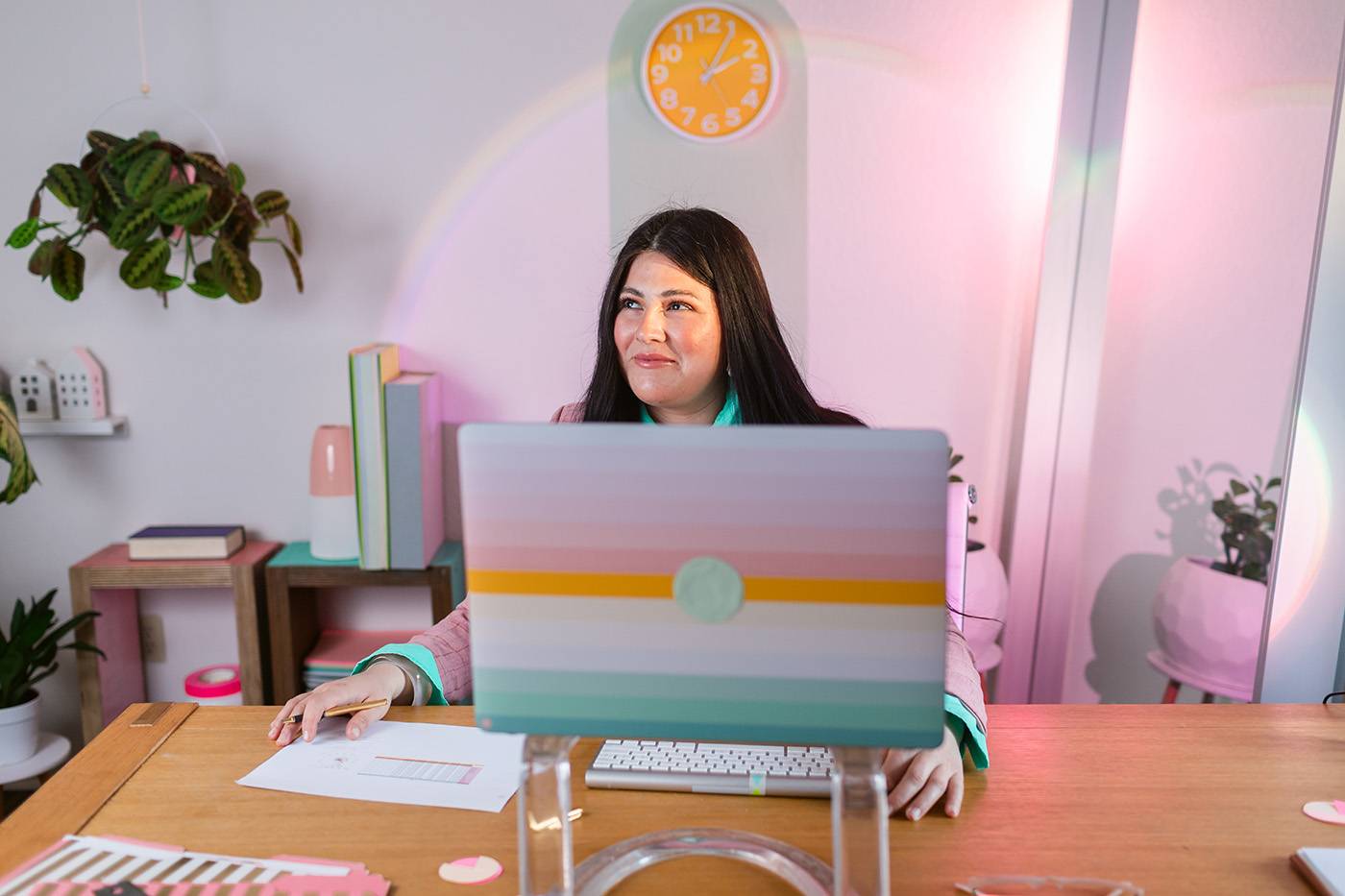 Female Entrepreneur in pink LED lights office working in a laptop on stand by graphics, rainbow and wall clock