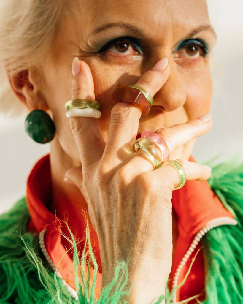 Close-up portrait of aged female covering her face with her hands, which are wearing many multi-colored fashionable rings. She looks up to the side and is wearing a green and red bow.
