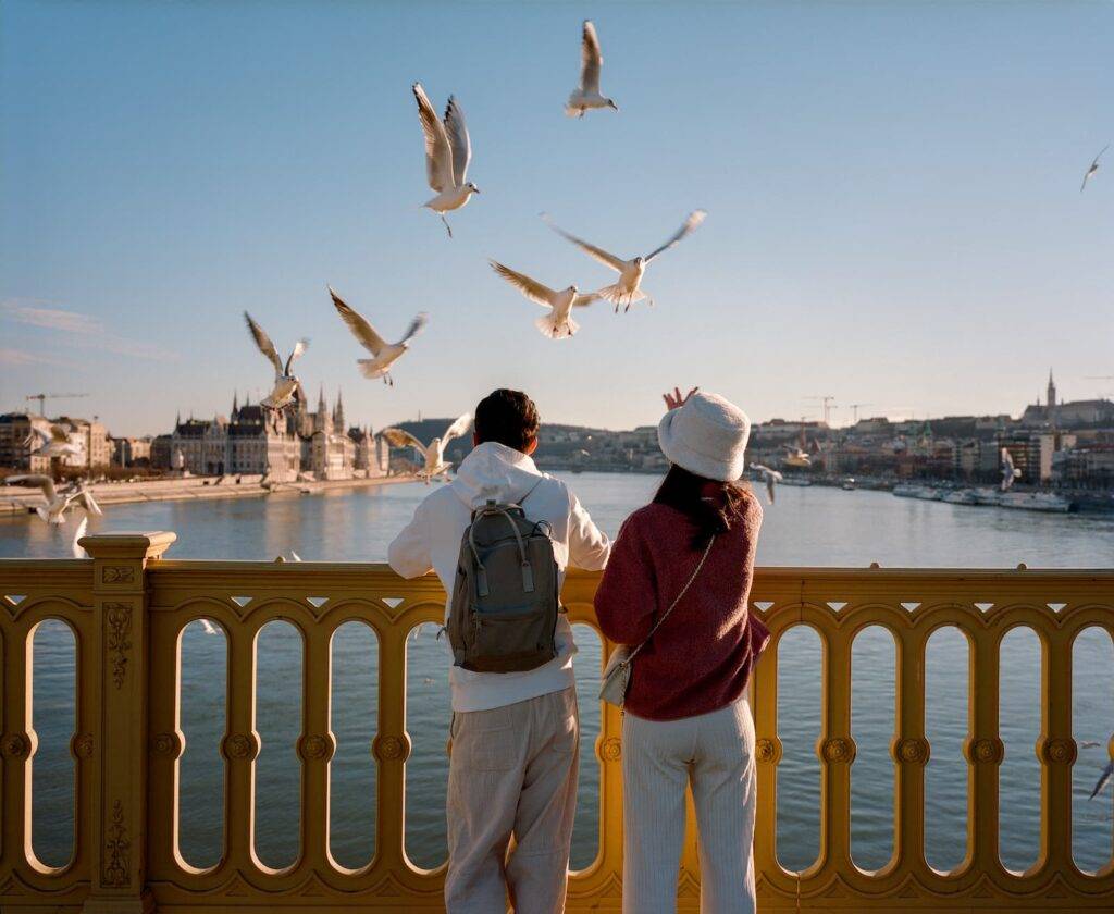A couple of tourists on a bridge in Budapest feeds seagulls against the background of the city and the Danube River