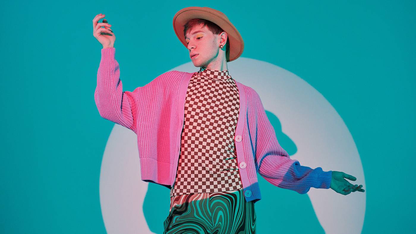 Horizontal studio portrait of stylish young gen Z queer Caucasian man wearing casual clothes with skirt and hat posing on camera in spotlight, blue background