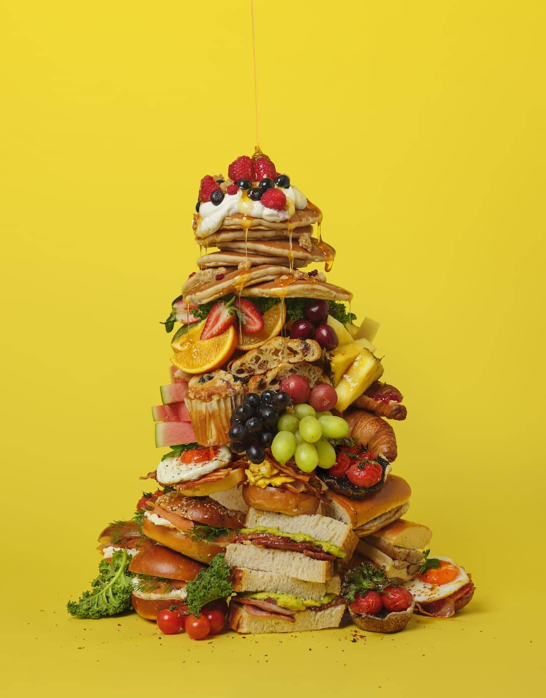 A huge pile of food is stacked up against a yellow isolated background. We see sandwiches, pastries, fruit, pancakes, waffles, syrup etc