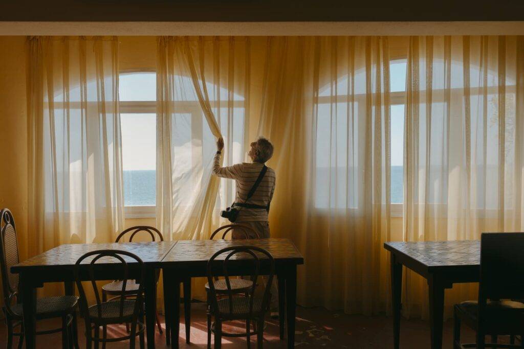 Man in empty restaurant with big windows with view of the Mediterranean Sea. The windows are covered by lace curtains creating a warm light. He's opening the curtains.