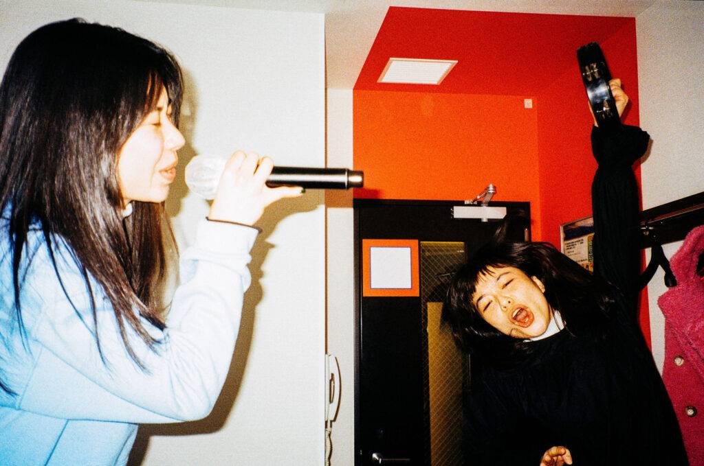 Two young women enjoy singing together in a karaoke booth. One is singing passionately into a microphone with closed eyes, the other is jumping holding a tambourine.