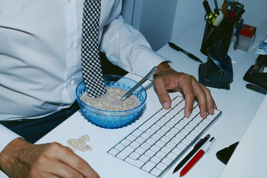 a man, wearing shirt and necktie, is having breakfast sitting at his office desk and is soaking accidentally his necktie in the bowl with overnight oats