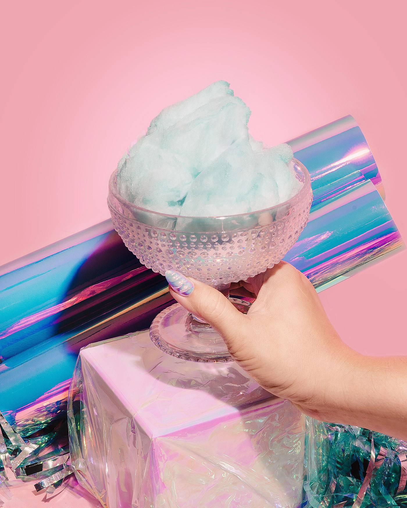 Iridescent ice cream glass holding blue cotton candy with blue sparkling streamers and holographic paper rolls. One caucasian hand holding glass with mermaid nail.