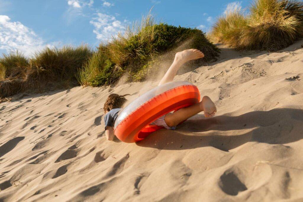 Barefoot anonymous boy at the beach having fun rolling down the dune with a red floatie during summer vacation