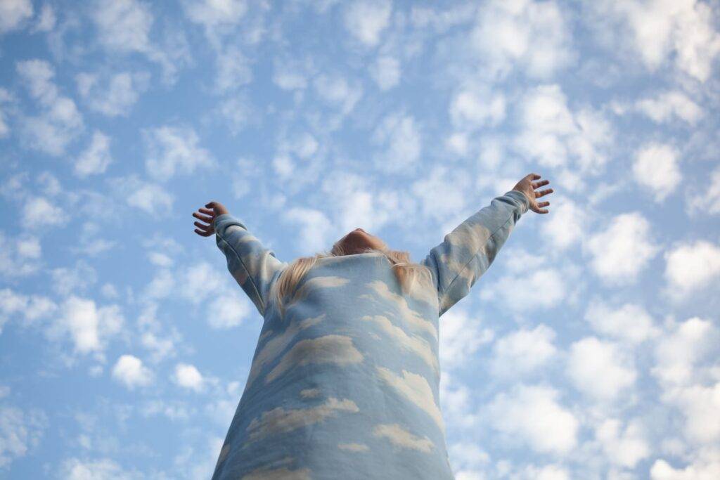 A child wearing a top covered in clouds stretches up into a vast blue sky. She is anonymous but her blonde hair is visible,