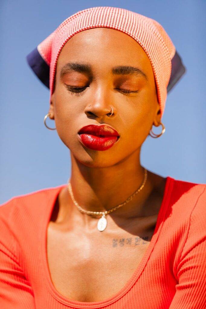 A portrait of a black woman stands on the sky background, wearing a red shirt, a red bandana, and red makeup