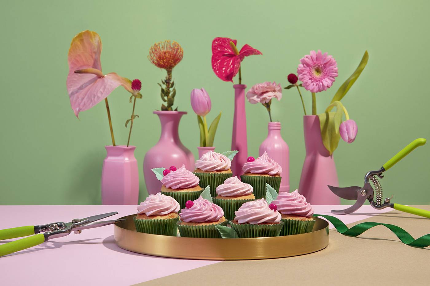A golden platter full of pink and green cupcakes that look like flowers. The platter is on a cute flower shop, next to gardening tools and in front of six pink vases full of flowers. The wall is green ant the table is pink.