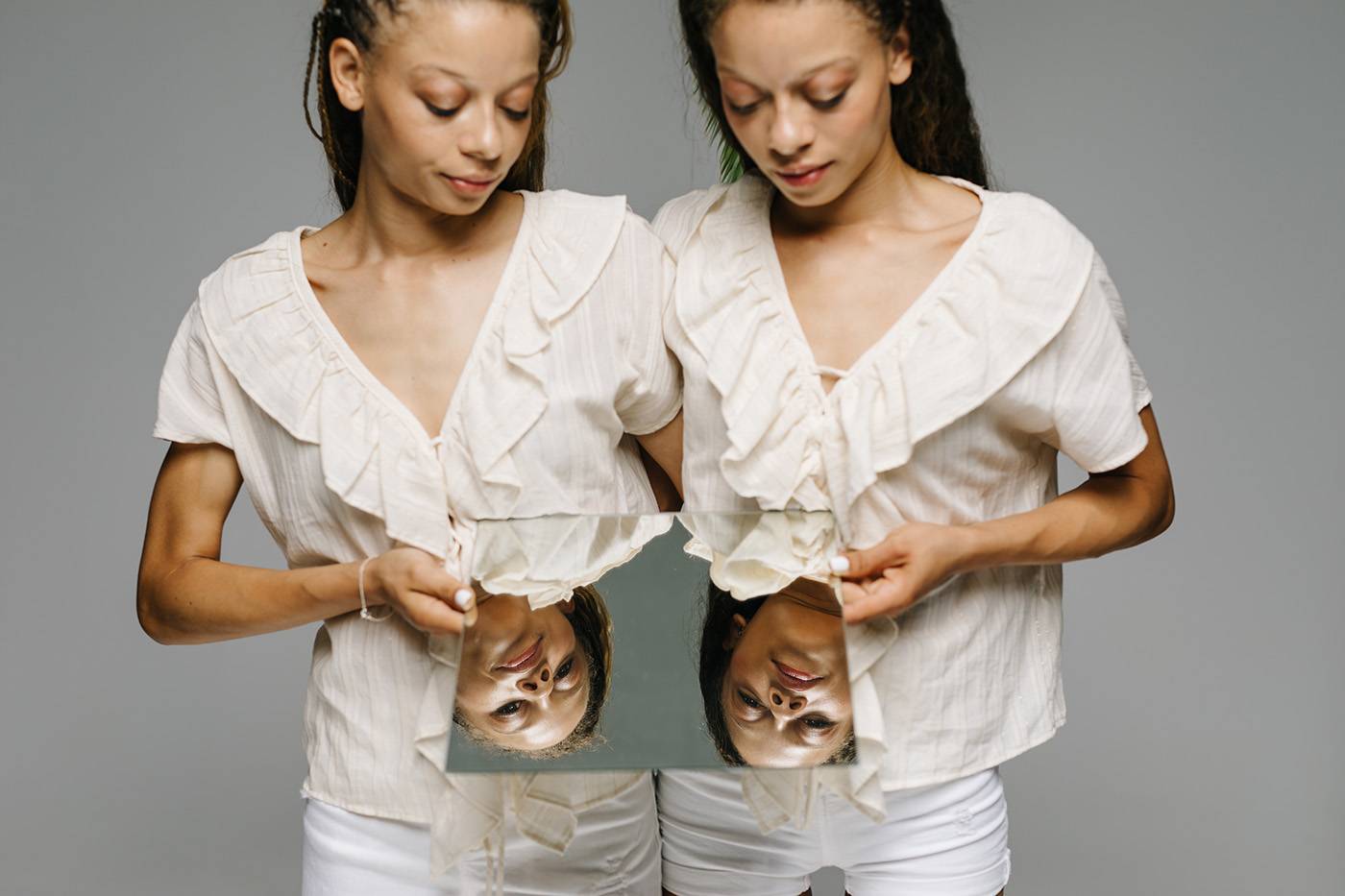 Two lovely young women in similar clothes†embracing each other and looking at reflection in mirror while standing on gray background