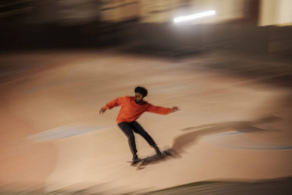 High angle man in casual clothes stretching out arms and riding longboard fast in evening in skatepark.