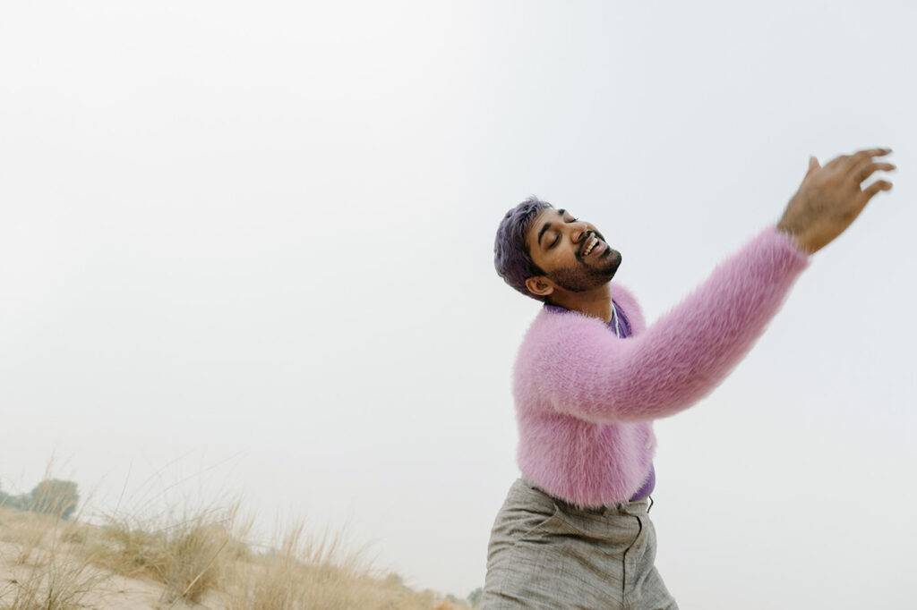 A young man with beard and purple hair smiling and dancing in the wilderness. A foggy day in the morning. He wears purple fluffy top, violet top and a pearl necklace.