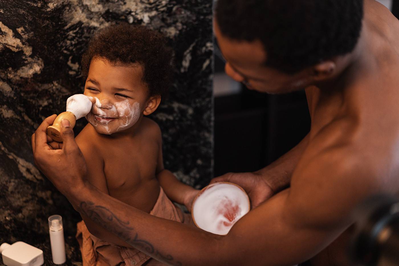 Father and son playing in the bathroom. Father is applying a shaving foam to a smiling kid's face.
