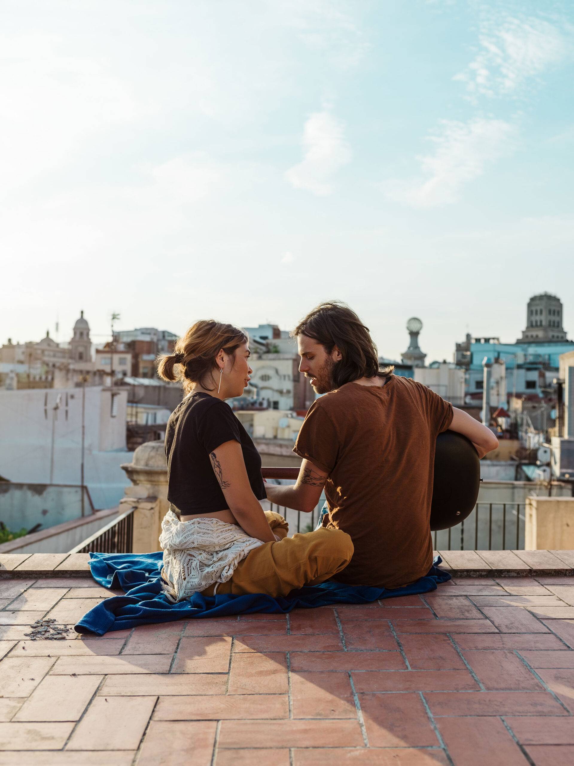 Back view of man with guitar witting on rooftop with stylish woman singing together on background of city and blue sky. Romantic dating