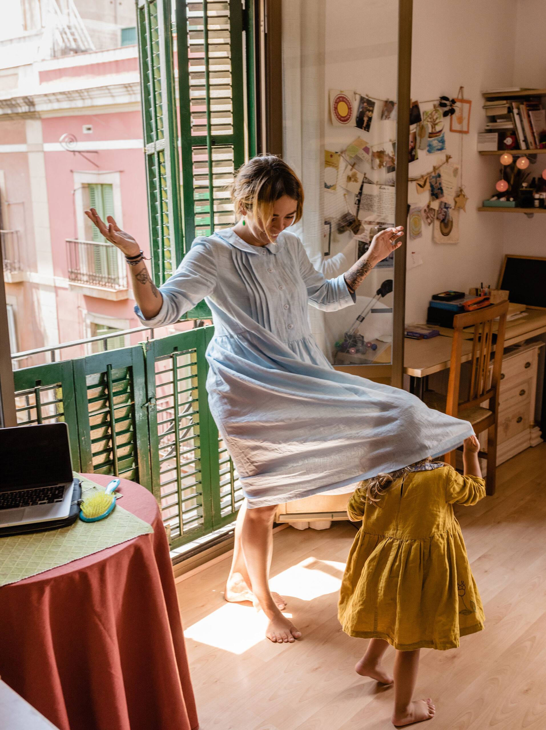 Adult cheerful woman in living room at home with little playful girl covering under skirt of dress having fun