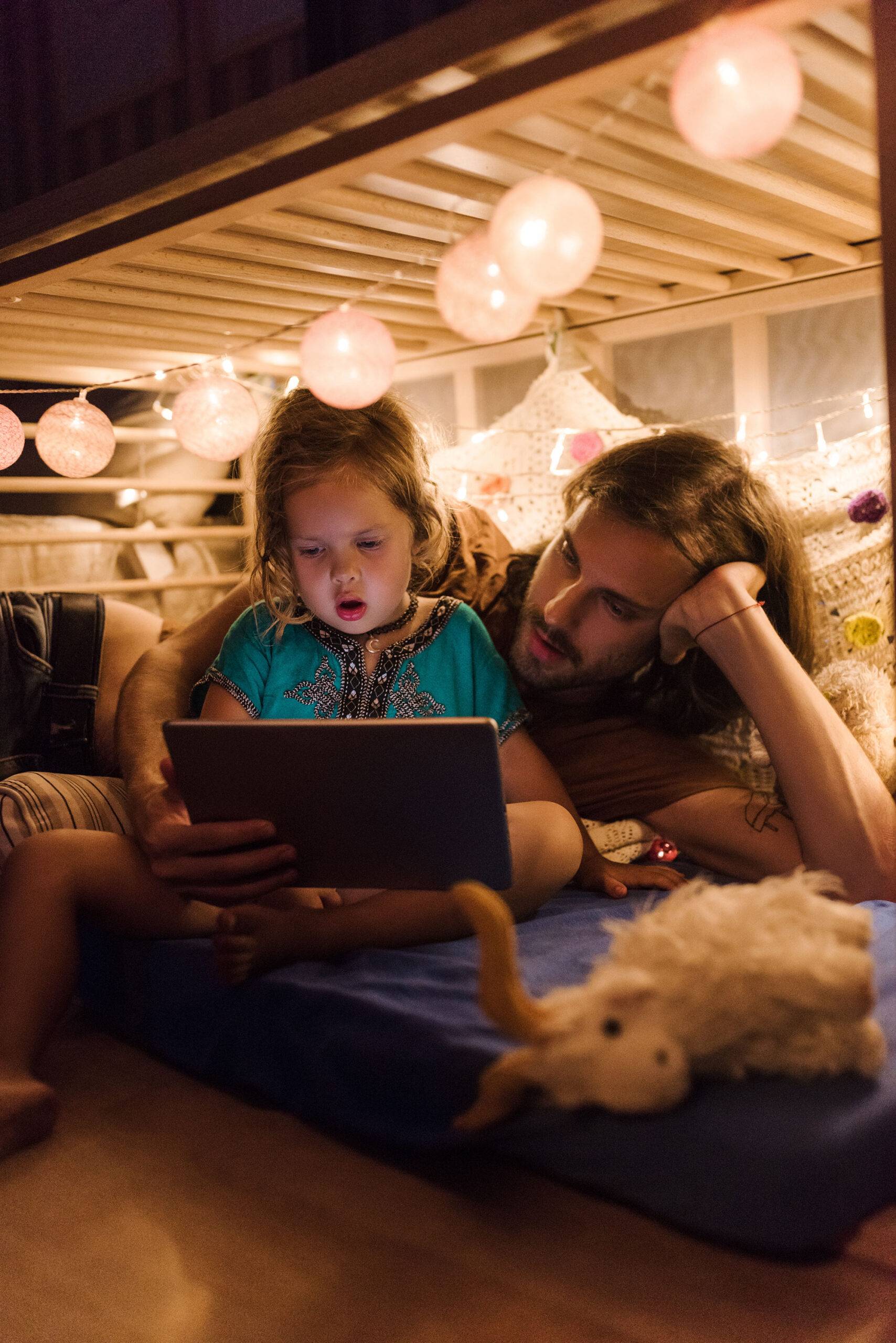 Little charming girl with bearded man cuddling on small cozy bed with garlands and watching cartoon on tablet together