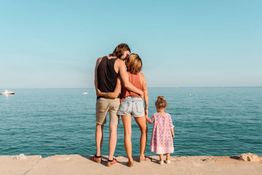 Back view of content happy couple kissing on waterfront standing with little girl on seascape background in sunlight
