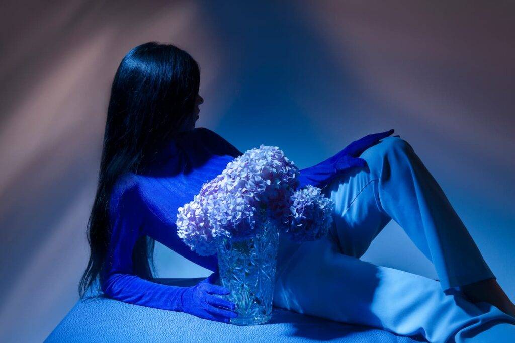 A close up of an anonymous female person with long hair dressed in a fashionable blue bodysuit posing in the darkness with flowers
