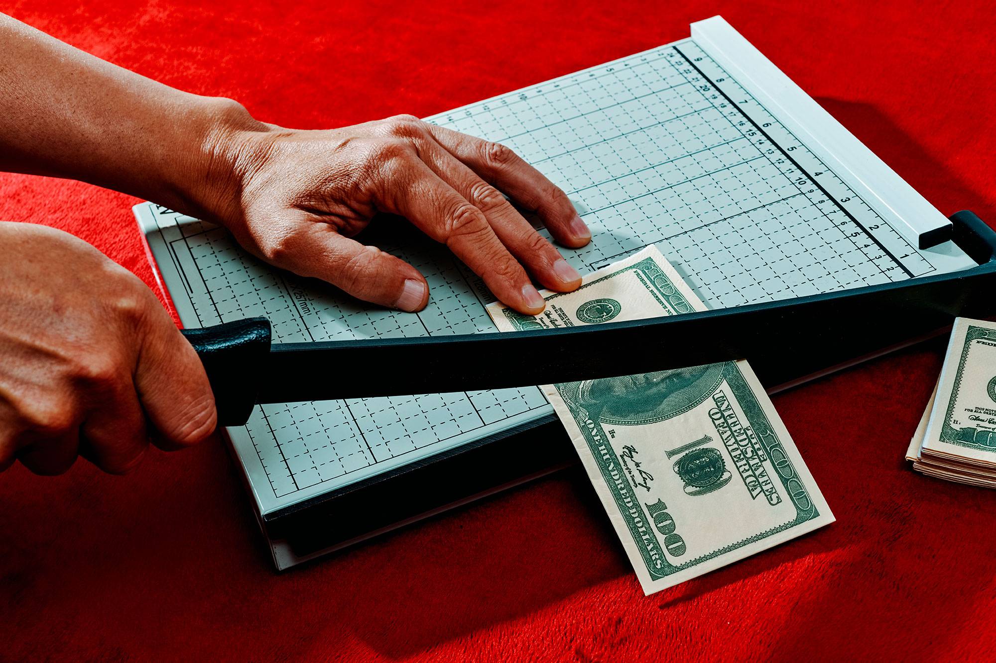 a man is cutting a fake 100 dollar baknote with a paper cutter placed on a fuzzy red fabric surface