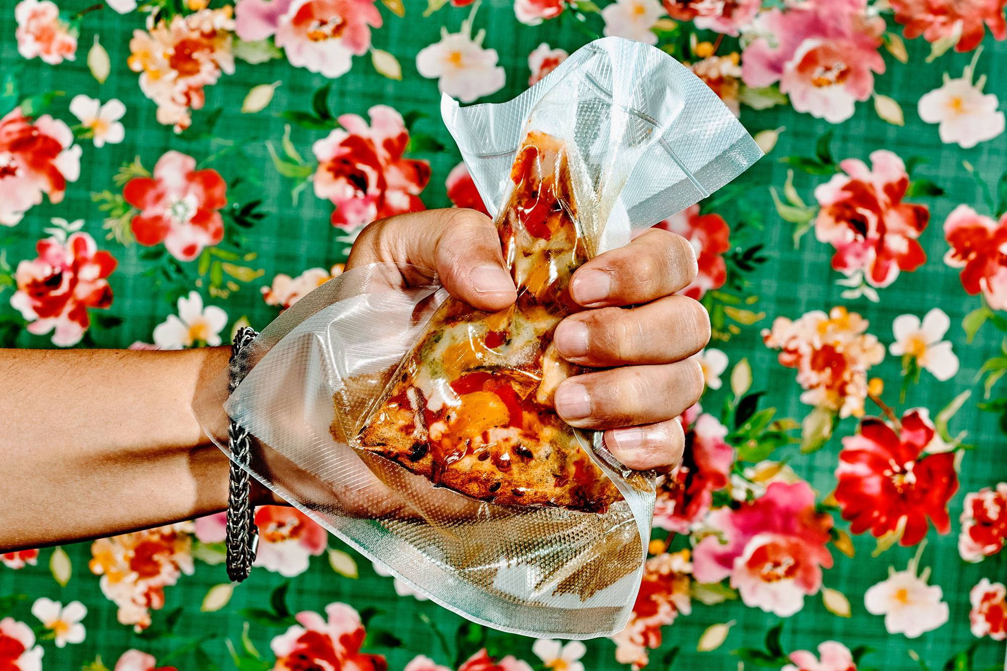 a man is grabbing tightly a vacuum packed slice of vegan pizza, in front of a floral-patterned wallpapered wall