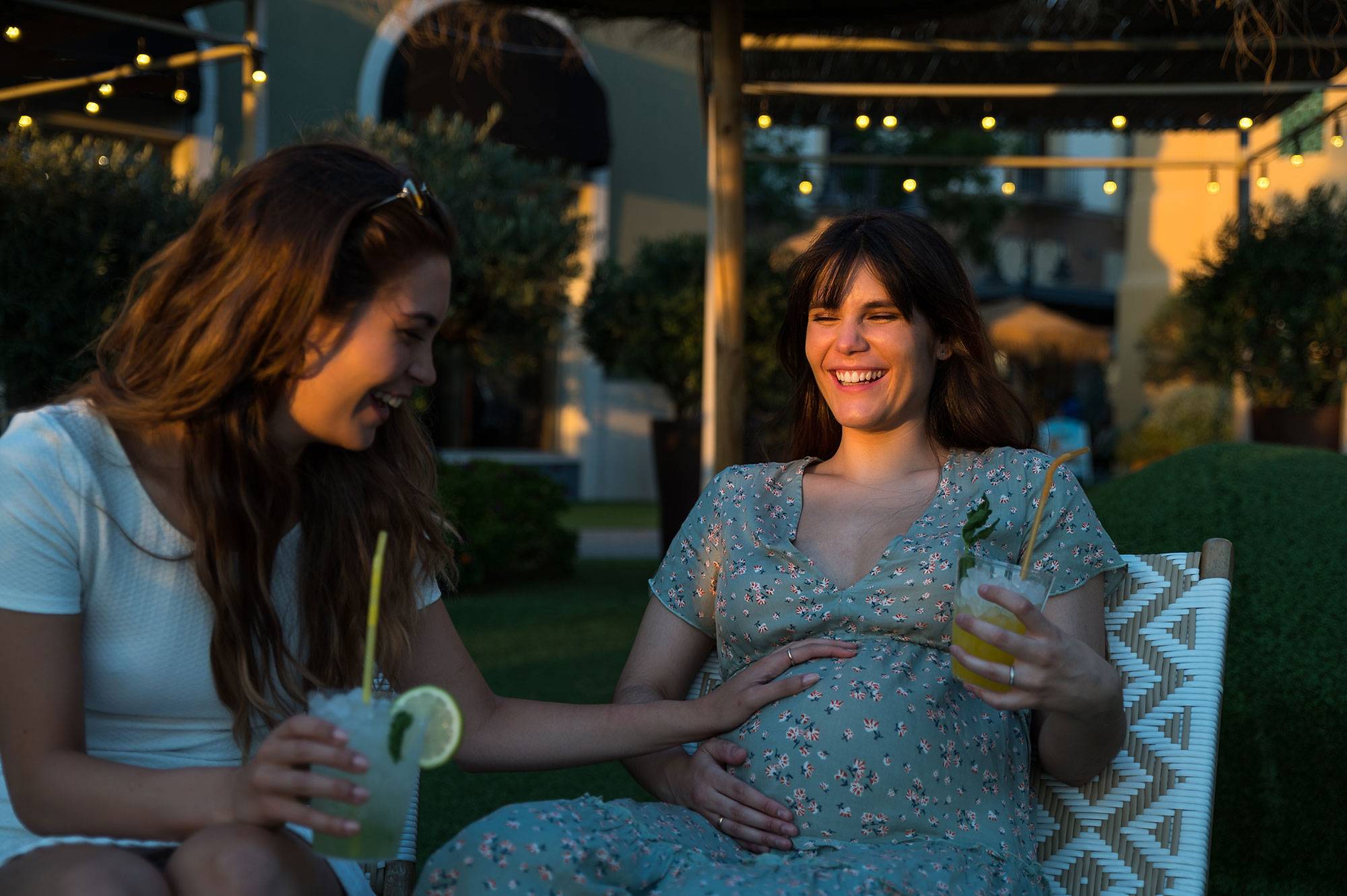 girlfriend with cold drink smiling and touching tummy of pregnant friend while resting on terrace together