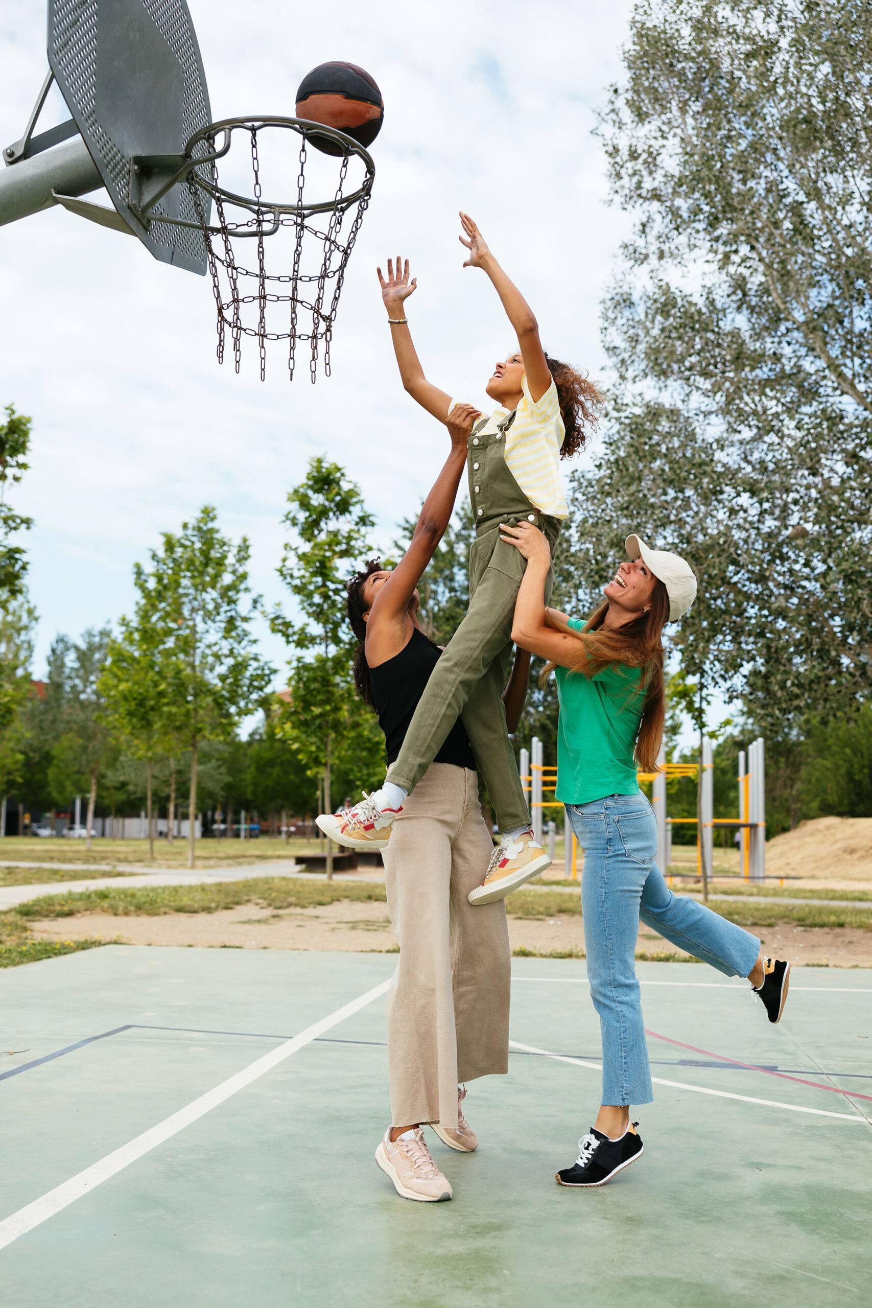Same-sex parents with their daughter playing basketball in outdoor basketball court on a summer day
