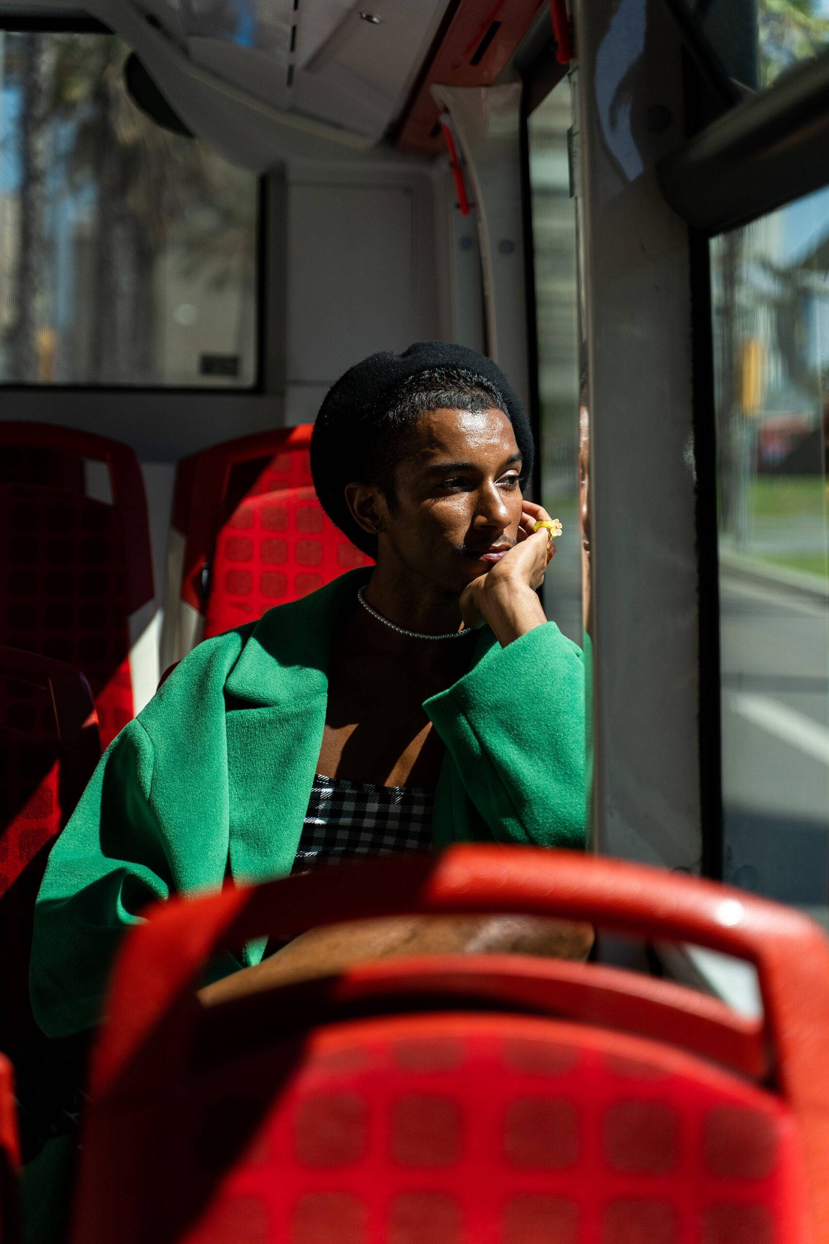 Pensive gender queer attractive african american man in green overcoat and beret looking out the window traveling by bus with red seats around the city