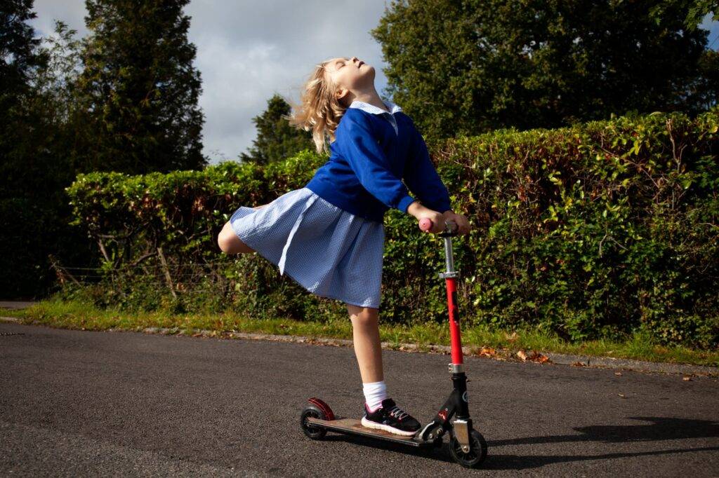 A little girl raises her face to the sunshine and her leg up behind her as she scoots down a road.