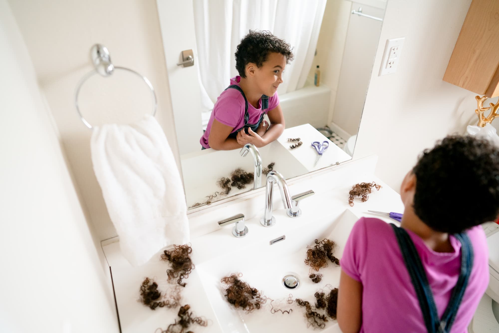 Girl with short, curly hair admires herself in the mirror. The counter in front of her is covered in dark curls and a pair of purple handled scissors rest nearby. Home haircut.