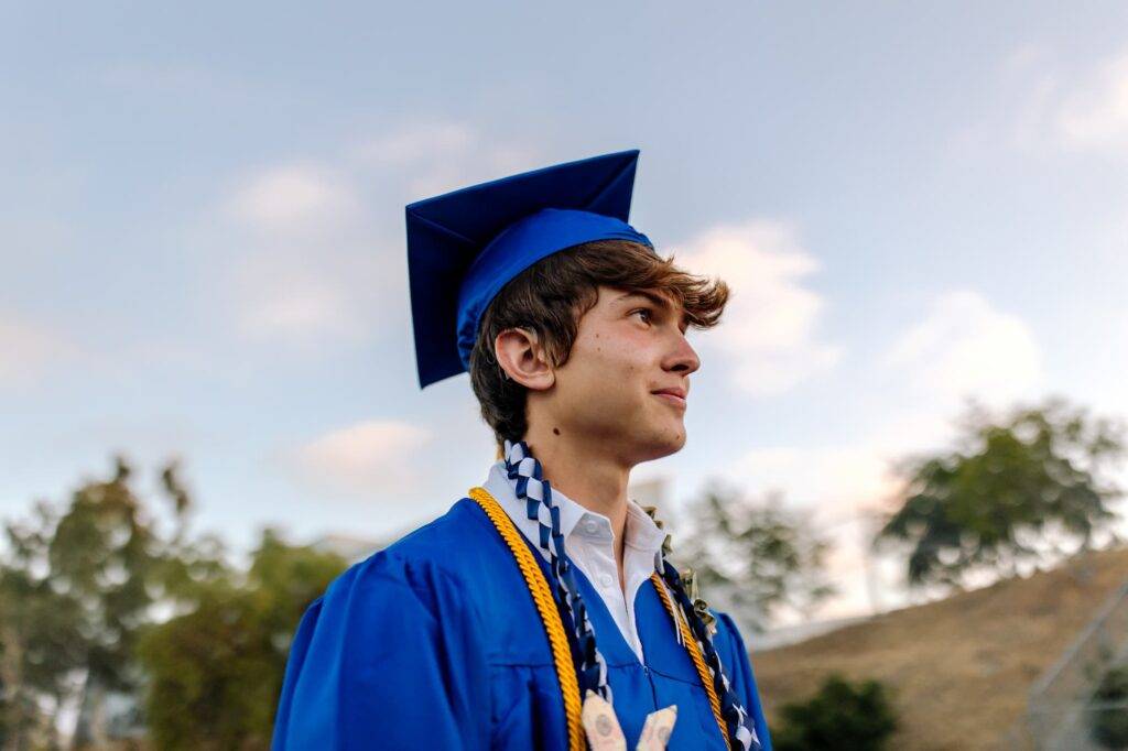 Young teen male with cochlear implant attends graduation ceremony. He is standing in front of blue sky with clouds wearing cap, gown, money lei, and tassel.