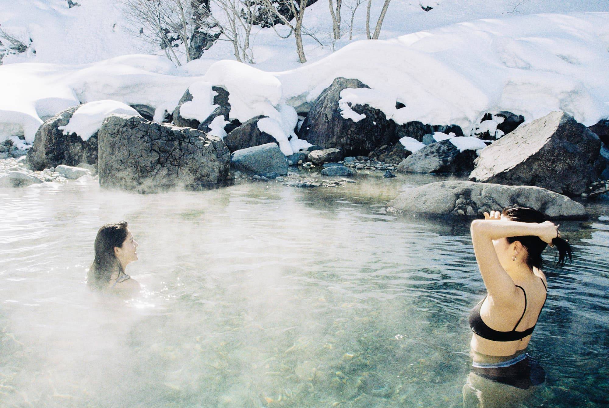Two slim women soak in a natural hot spring in the winter. Behind them are rocks and trees covered in snow.