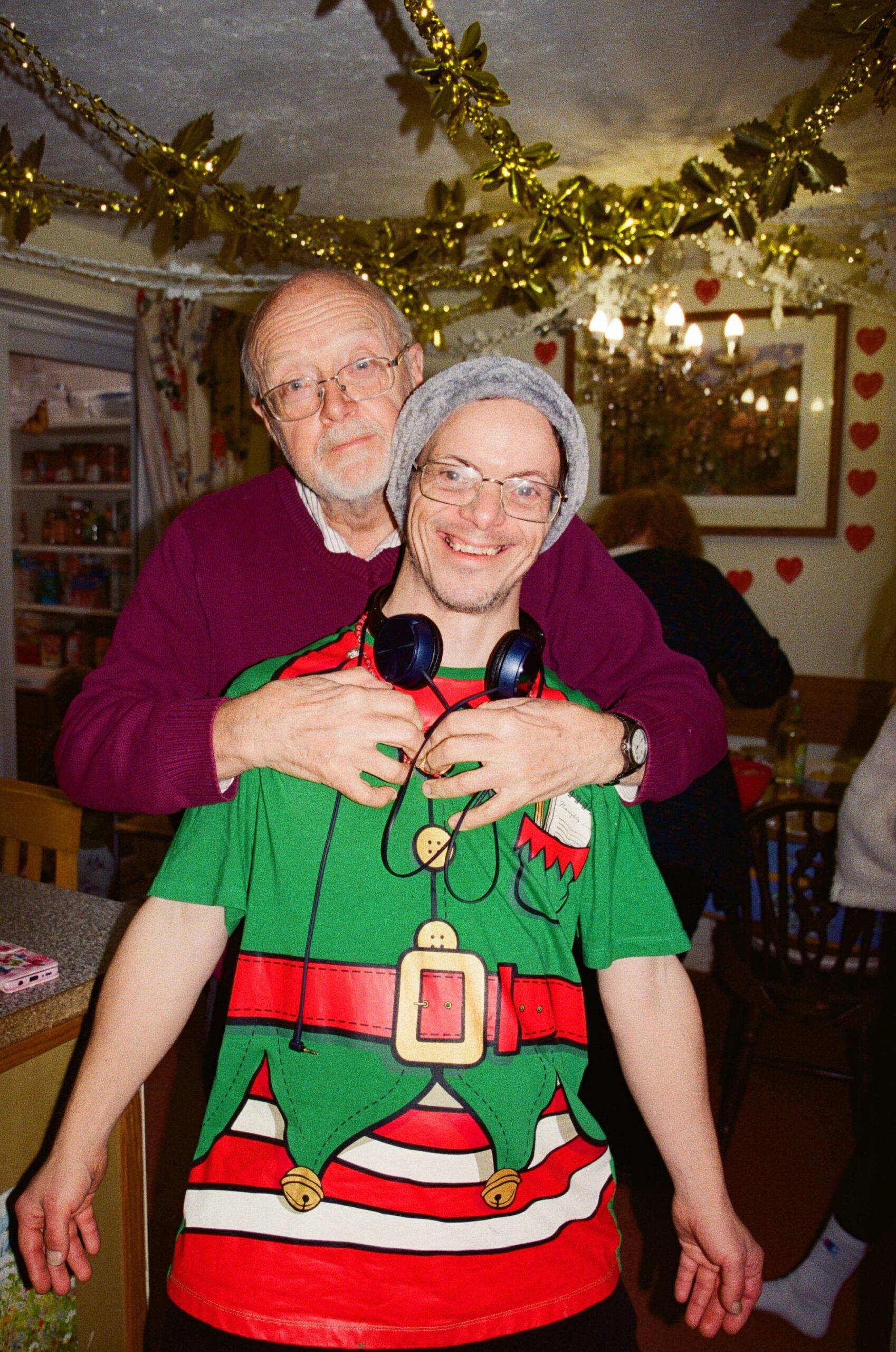 Father And Son Share A Moment At Christmas A man with downs syndrome smiles broadly at the camera while his father hugs him. They are in a dining room decorated for Christmas. Film image with flash.