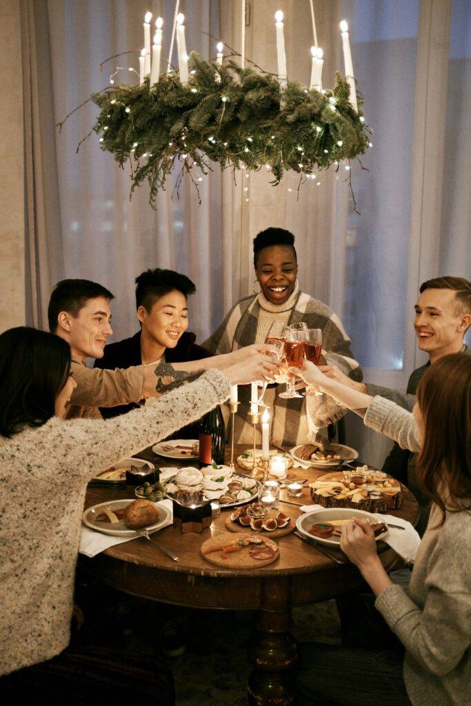 Multiethnic group of young people clinking glasses with champagne while sitting at holiday table in beautifully decorated apartment