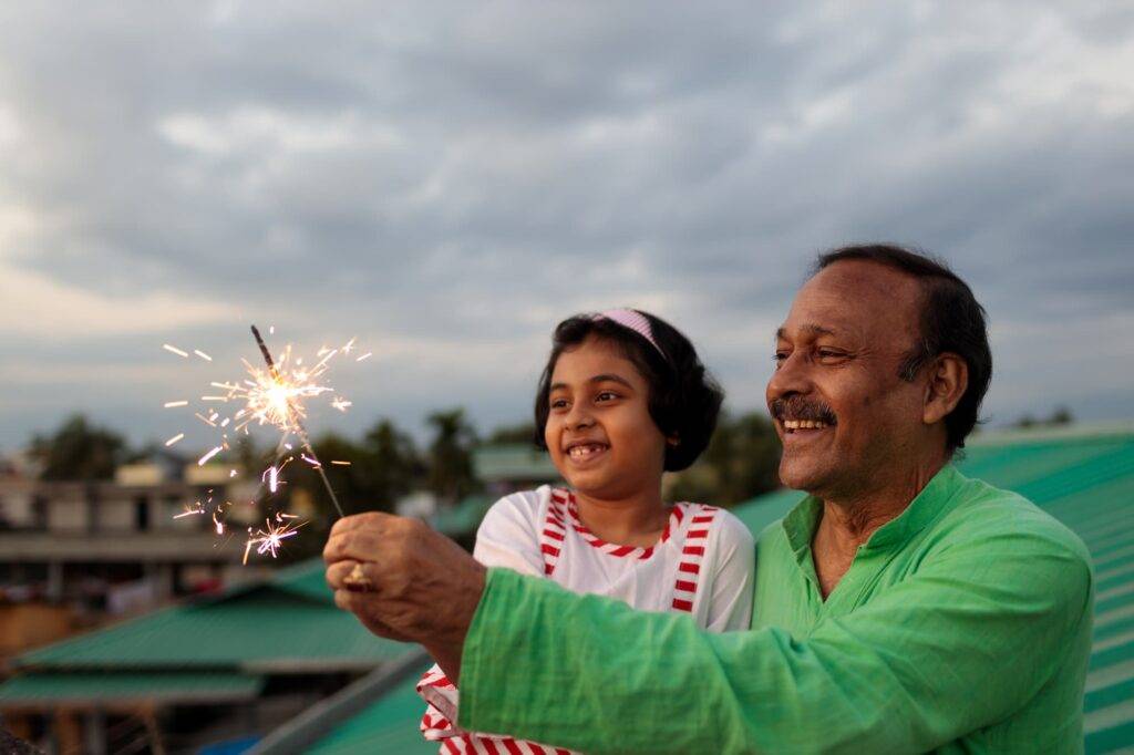 Little Girl And Her Grandfather Having Fun With Sparklers Outdoors