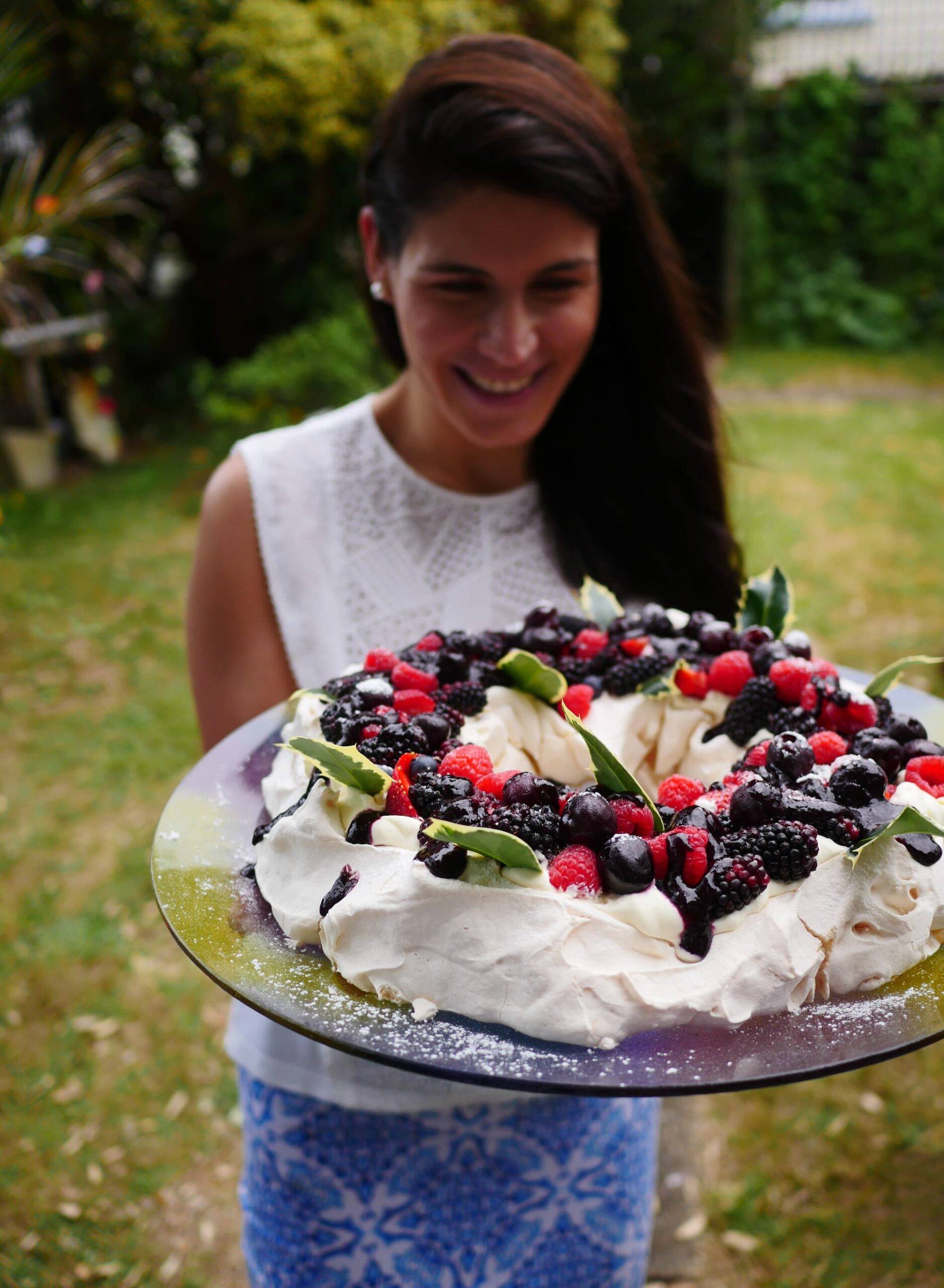 A slender, healthy woman holding a huge pavlova cake. The meringue cake is topped with fresh berries, whipped cream and holly leaves. The photo was made at christmas, which is the middle of summer in New Zealand.