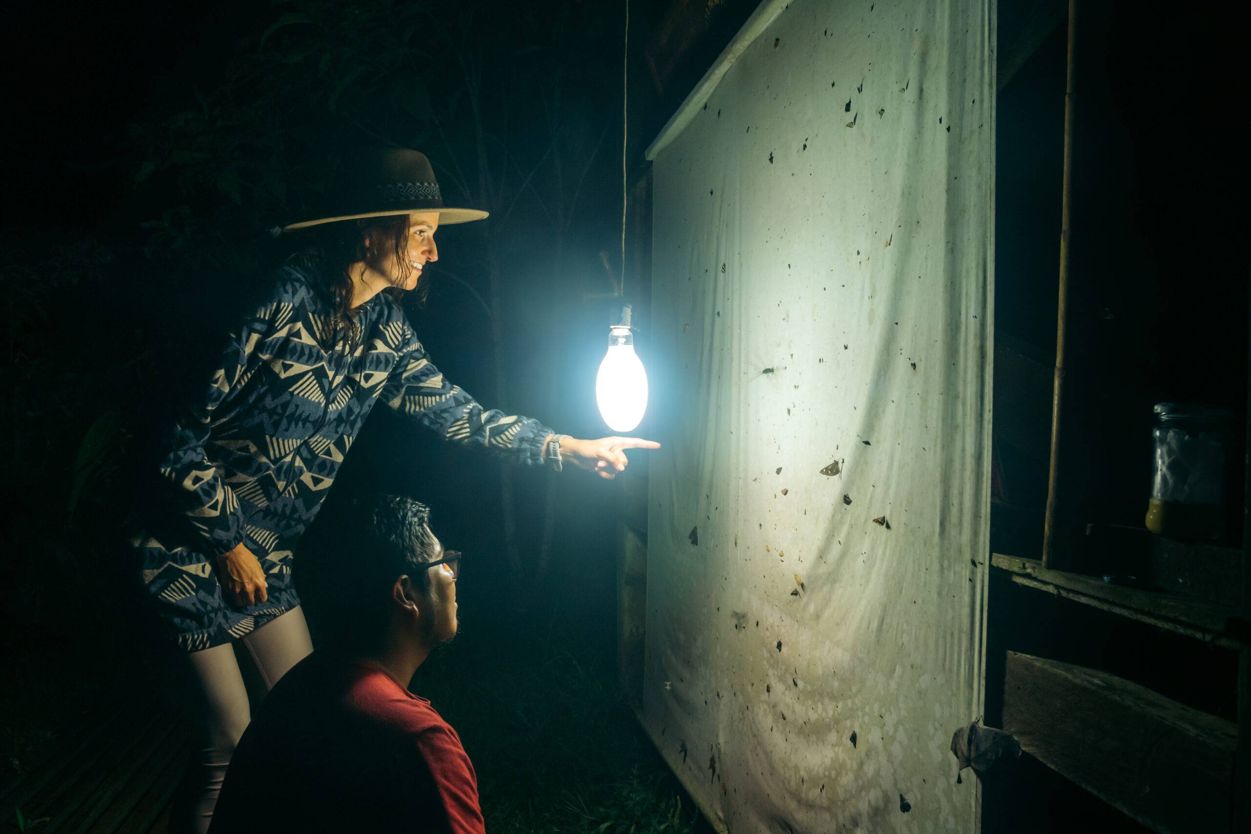 White woman with traveler clothes and latin man smiling while doing conservation work at night with a light trap for insects. They are doing ecotourism activities in the Peruvian Amazon Jungle.
