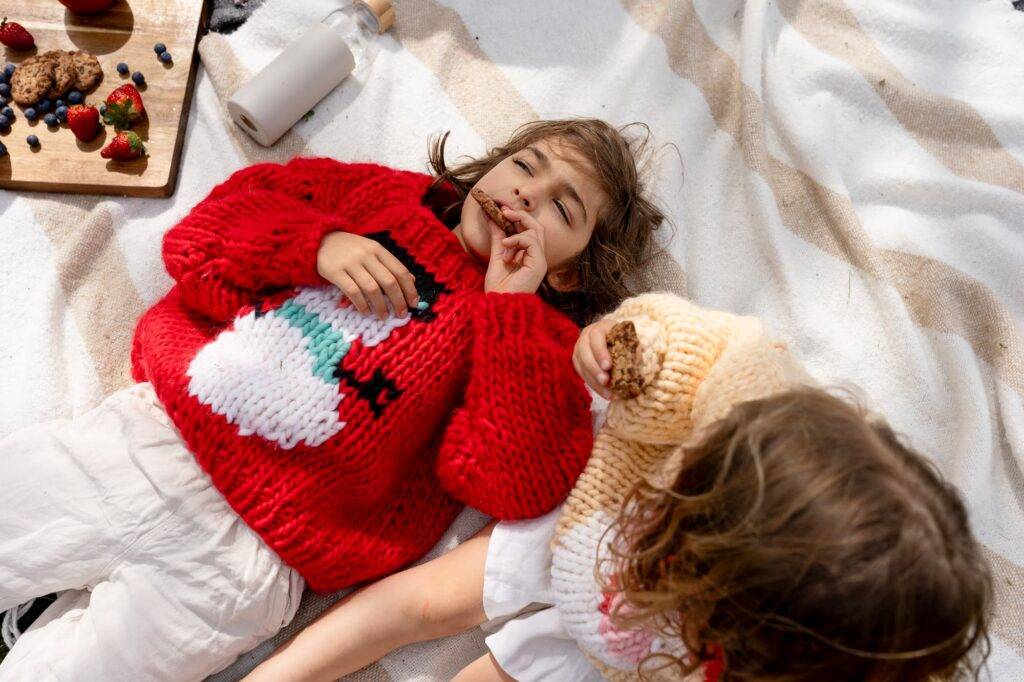 Kids In Christmas Knits Having A Picnic And Playing With Sone Cookies.