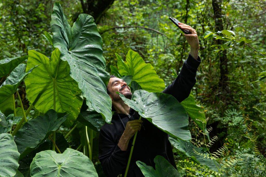 A man with beard taking a selfie in a forest surrounded by huge green leaves