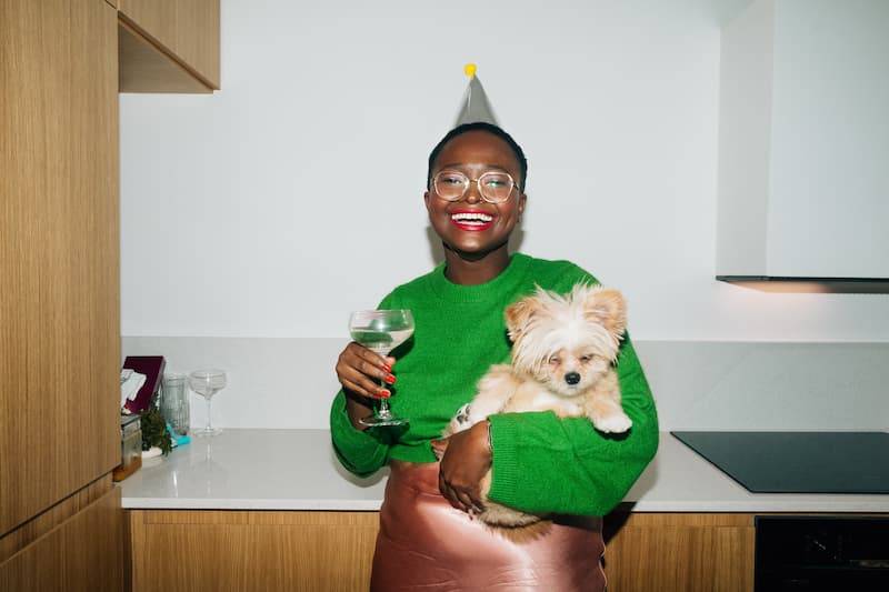Beautiful young woman drinking champagne, holding cute little dog and looking at camera in kitchen at house Christmas party