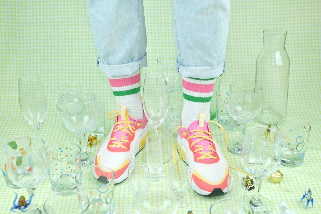 The legs of an unrecognisable person standing among fragile objects - glasses and toys made of glass. On the person's feet are sneakers, one of which is untied
