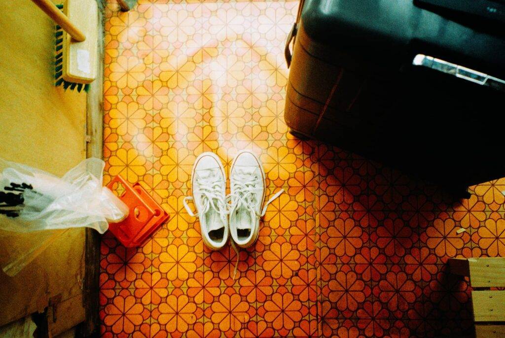 A house entrance with retro, orange linoleum. A large black suitcase, white sneakers, clear plastic umbrella and a broom are seen in the photo. Taken from above.