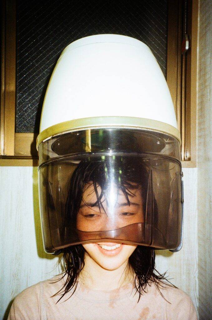 A smiling Japanese woman uses a retro bonnet style hair dryer on her wet hair.