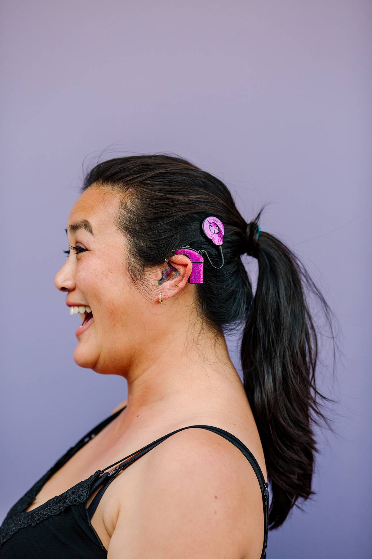 Profile of smiling Asian female in black camisole with hair in ponytail. She has a disability and wears hearing device. Purple wall in background.