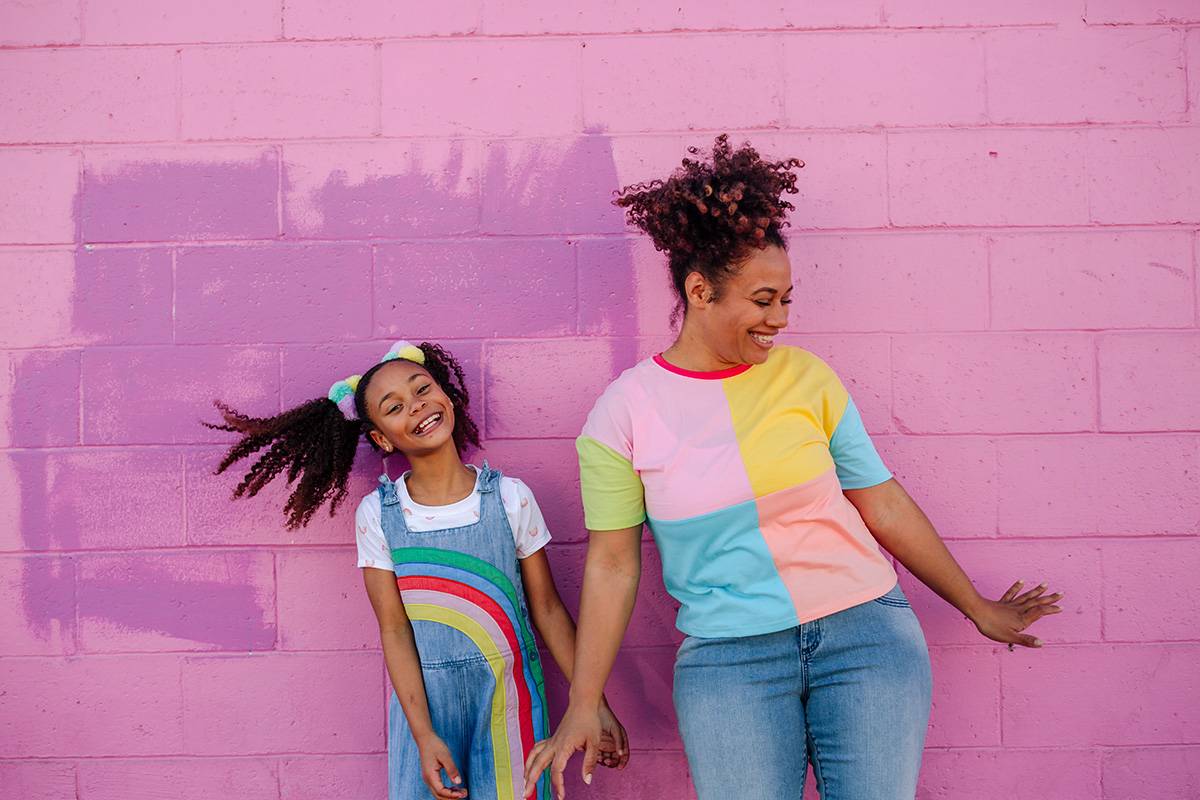 Mixed race daughter in rainbow overalls standing with curvy mother in jeans and color-block shirt against pink painted block wall.
