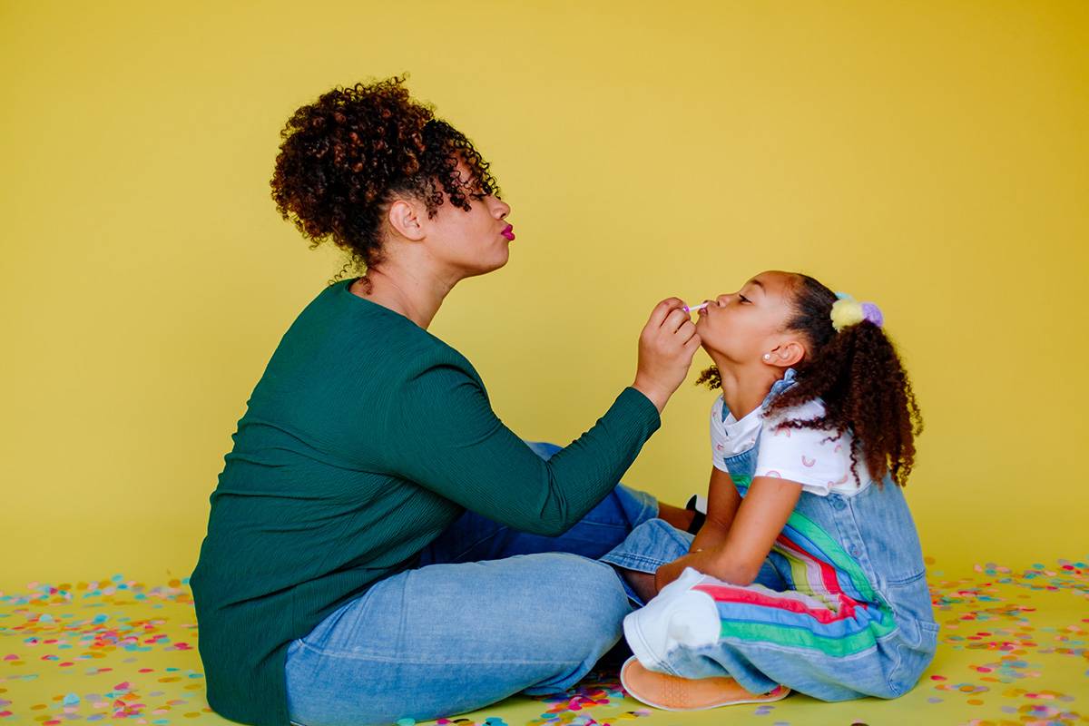 Profile of beautiful mother applying lipstick to sweet daughter with pigtails while sitting in confetti. Yellow studio background.