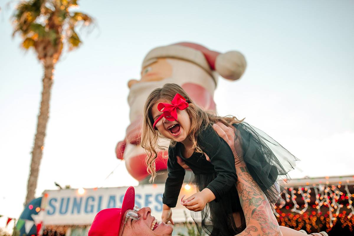 Smiling Father lifts joyful daughter into air at Christmas Tree Lot. Inflatable Santa and palm tree in background. Girl is wearing red bow and black dress.